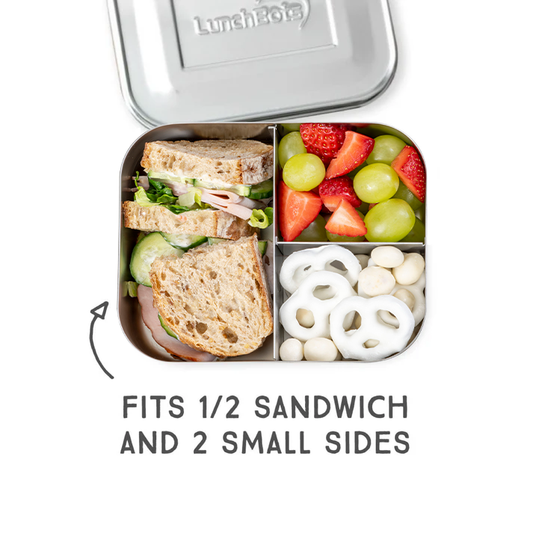 A silver LunchFits stainless steel lunch box shown open with a sandwich, fruit, and small sides in the compartments. Text below reads 'FITS 1/2 SANDWICH AND 2 SMALL SIDES' with a curved arrow pointing to the box.